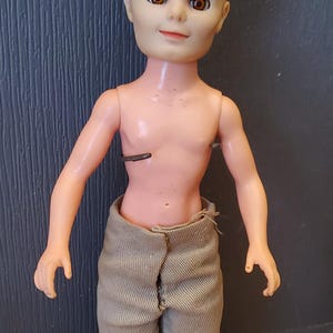 May include: A plastic doll with blonde hair, brown eyes, and a light brown shirt. The doll is standing on a grey base.