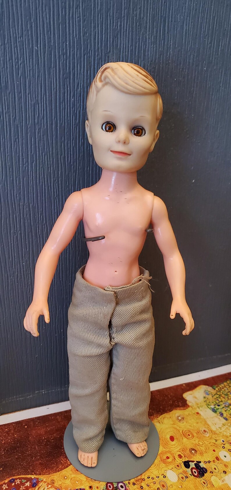 May include: A plastic doll with blonde hair, brown eyes, and a light brown shirt. The doll is standing on a grey base.