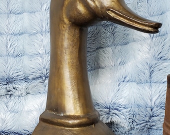May include: A gold-colored metal sculpture of a duck head with a long neck and a base. The sculpture is on a blue and gold patterned fabric.