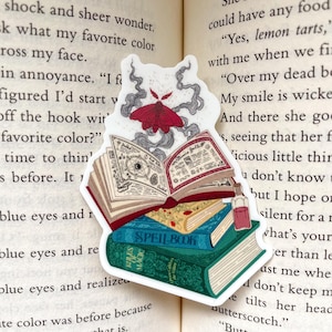 May include: A sticker featuring a stack of three books with a red moth flying above them. The books are open and have pages filled with handwritten text. The top book has the title "SPELL BOOK" written on the spine. The sticker is on a page of a book with text.