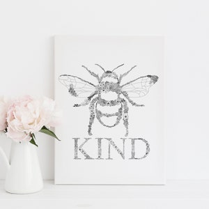 May include: A black and white print of a bee made of flowers with the word "KIND" underneath. The print is framed and sitting on a white shelf with a white pitcher and pink flowers.