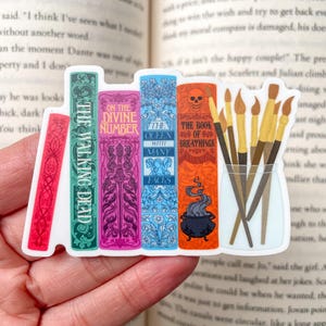 Könnte beinhalten: Ein Aufkleber mit einem Stapel von fünf bunten Büchern mit Titeln wie "The Walking Dead", "On the Divine Number", "The Queen with Many Faces", "The Book of Breathings" und einem Kessel, aus dem Rauch aufsteigt. Außerdem sind fünf Pinsel in einem Glasgefäß zu sehen.