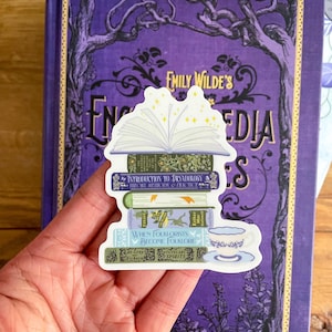 May include: A sticker of a stack of books with a magical open book on top. The books have titles like "Introduction to Dryadology", "When Folklorists Become Folklore", and "Tutelary Spirits". There is a small teacup and saucer next to the books.