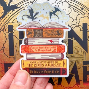 May include: A stack of four books with dragon-themed covers. The top book is open, revealing a white page with three black dragons flying above it. The book titles are "Fables of the Barren", "Colonel Kaori's Field Guide to Dragonkind", "Major Afrenda's Guide to the Riders Quadrant", and "The Dragon Riders Codex".
