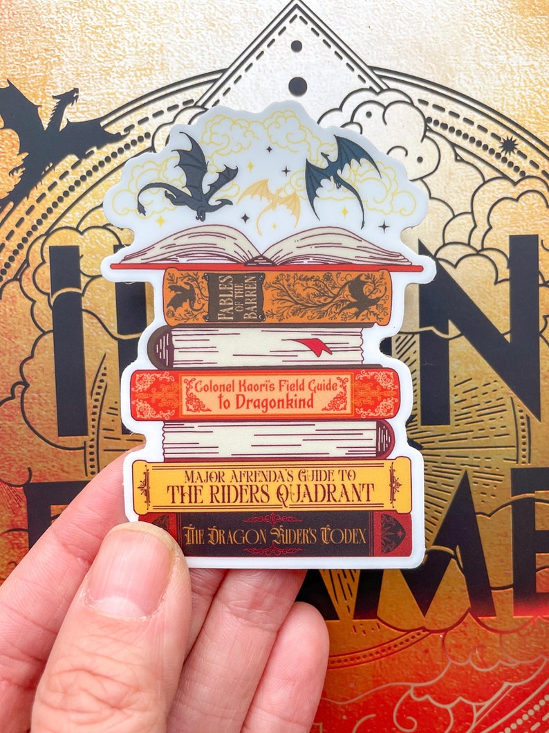 May include: A stack of four books with dragon-themed covers. The top book is open, revealing a white page with three black dragons flying above it. The book titles are "Fables of the Barren", "Colonel Kaori's Field Guide to Dragonkind", "Major Afrenda's Guide to the Riders Quadrant", and "The Dragon Riders Codex".
