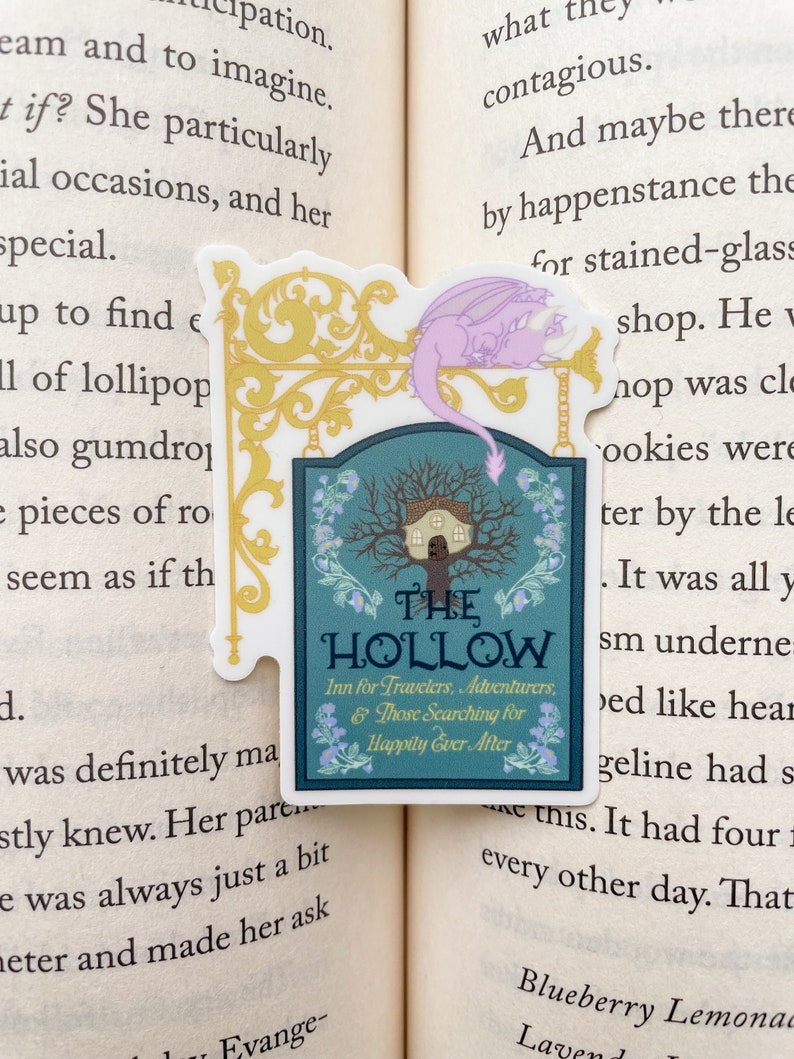 May include: A sticker featuring a sign for "The Hollow Inn for Travelers, Adventurers, & Those Searching for Happily Ever After". The sign is decorated with a whimsical tree and a small house. A purple dragon sticker is placed above the sign.