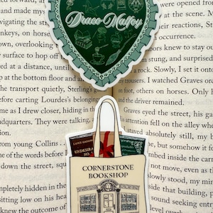 Pode incluir: Dois adesivos em uma página de livro. Um é um adesivo verde em forma de coração com as palavras "Draco Malfoy" e ilustrações. O outro é um adesivo de sacola bege com as palavras "Cornerstone Bookshop" e um desenho de uma loja.