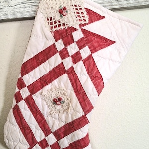 May include: A quilted Christmas stocking featuring a red and white woven-style pattern. The stocking is accented with cream-colored crocheted lace patches, each centered with small embroidered red berries and green leaves, and includes a hanging loop.