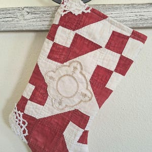 May include: A quilted Christmas stocking featuring a red and white geometric patchwork pattern. The stocking is accented with white crocheted lace trim at the top and bottom edges and features a central circular embroidered applique detail.