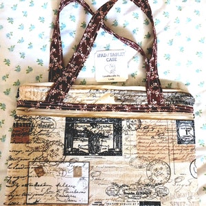 May include: An iPad/tablet case with a vintage postcard design. The beige fabric is covered in French script, stamps, and illustrations. It has a zipper closure and brown floral handles. The tag reads "IPAD/TABLET CASE" and "Lovablecrafts by Linda."