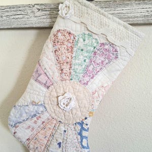 Op de afbeelding: Een gewatteerde kerstkous met een patchwork waaierontwerp in diverse pastelkleurige bloemen- en patroonstoffen. De kous is versierd met witte gehaakte bloemen, een witte kanten rand aan de bovenkant en een witte lus om op te hangen.
