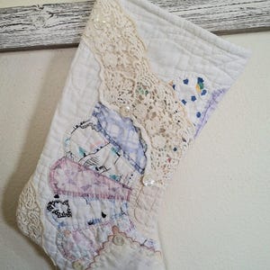 Pode incluir: Uma meia de Natal acolchoada com um design de patchwork de tecido branco, padrões florais e estampas de rosas azuis. A meia é adornada com uma borda de renda cor de creme e vários pequenos botões brancos na superfície frontal.