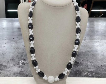 May include: A black and white beaded necklace displayed on a mannequin. The necklace features alternating black, oval-shaped beads and white, bone-shaped beads. A large, white, round bead is at the center. The necklace is a fashion accessory.