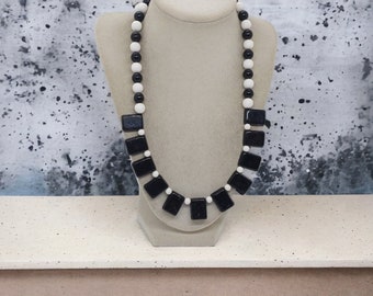 May include: A black and white beaded necklace with square and round beads. The necklace is displayed on a mannequin.