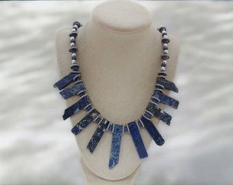May include: A blue and silver necklace with a pendant made of multiple blue and white stone pieces. The necklace is on a white mannequin.