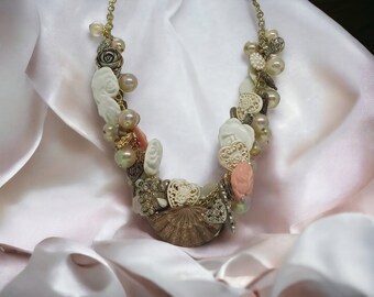 May include: A delicate necklace made of white and pink plastic flowers, gold chain, and pearl-like beads. The necklace is displayed on a pink satin fabric.