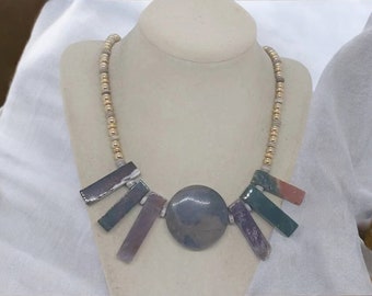 May include: A necklace with a large, round, gray and green stone pendant. The pendant is surrounded by smaller, rectangular stones in shades of purple, green, and brown. The necklace is made of gold and beige beads.
