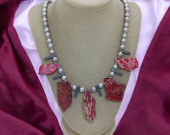 May include: A handcrafted necklace featuring a strand of gray faux pearls, accented with dark gray rectangular beads and irregularly shaped red jasper stones. The necklace is displayed on a mannequin against a backdrop of burgundy and pink fabric.