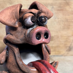 May include: A ceramic pig figurine with a large, open mouth and a long, red tongue sticking out. The pig has brown and pink coloring and large, round eyes.