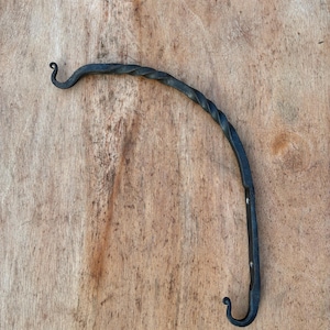 May include: A hand-forged, black metal plant hanger with a curved design. The top features a decorative spiral, and the bottom has a hook shape. The hanger is mounted on a wooden surface.