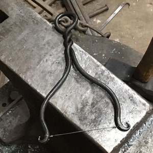 May include: A black metal blacksmith tool with a twisted handle and two hooks on the ends. The tool is resting on a metal anvil.