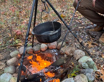 May include: A black cast iron cooking pot suspended over a campfire. The pot hangs from a black metal tripod. The fire is surrounded by stones and burning wood. The scene is set outdoors with autumn foliage.