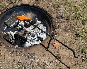 May include: A campfire scene with a metal fire pit filled with wood and ash. Two orange hot dogs are held over the fire by a long, black metal roasting stick. The ground around the pit is covered in dry grass and dirt.