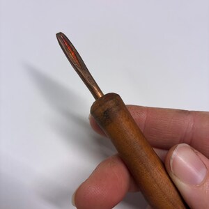 May include: A close-up of a wooden crochet hook with a copper-colored metal hook. The wooden handle is a warm brown color, and the hook has a pointed tip with a reddish hue.