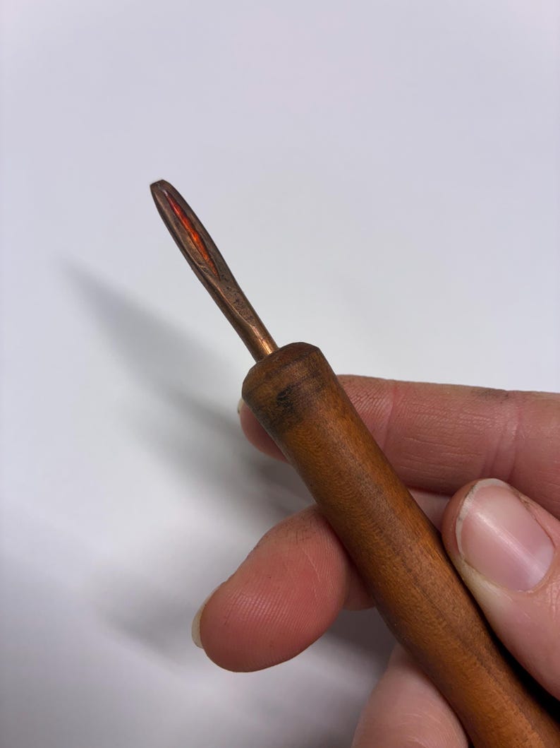 May include: A close-up of a wooden crochet hook with a copper-colored metal hook. The wooden handle is a warm brown color, and the hook has a pointed tip with a reddish hue.