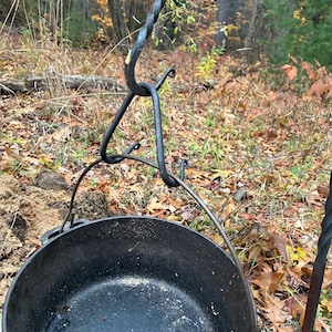 May include: A black cast iron Dutch oven pot hanging from a wrought iron tripod. The pot is round with a handle and sits over a fire pit. The tripod has decorative swirls and hooks. The background shows a forest with fall foliage.