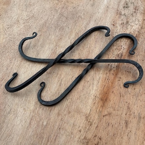May include: Two black, hand-forged iron hooks with a twisted central bar and curved ends. The hooks are designed for hanging items and are displayed on a light brown wooden surface. The hooks have a rustic, handcrafted appearance.