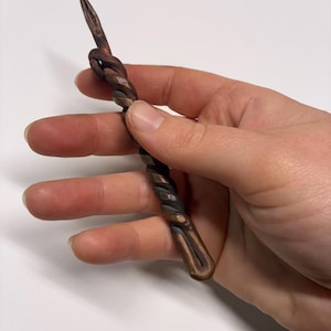 May include: A hand-forged, twisted metal tool with a pointed end and a looped knot. The metal has a dark, oxidized finish with copper highlights, creating a rustic, handcrafted appearance. The tool's design suggests it may be used for crafting or decorative purposes.