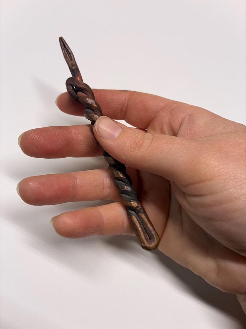 May include: A hand-forged, twisted metal tool with a pointed end and a looped knot. The metal has a dark, oxidized finish with copper highlights, creating a rustic, handcrafted appearance. The tool's design suggests it may be used for crafting or decorative purposes.