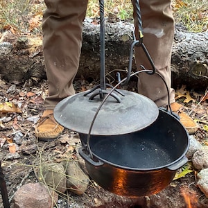 May include: A black cast iron Dutch oven with a lid, suspended over a campfire. The pot is hanging from a wrought iron support. The scene is outdoors, with a person in brown pants and moccasins visible in the background.