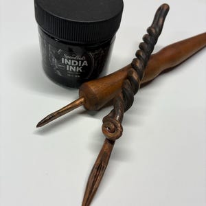 May include: A jar of Speedball India Ink with a black lid next to two wooden calligraphy pens. One pen has a twisted design, and the other is a simple, straight design. The pens are brown and the ink is black.