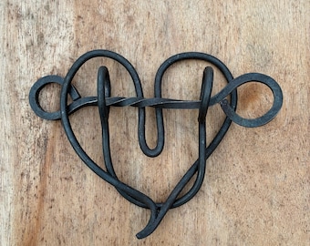 May include: A handcrafted, black metal heart-shaped sculpture. The sculpture is made of twisted metal rods, forming a continuous loop with a central, twisted section. The design includes two circular elements on either side of the heart.