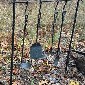May include: A black, hand-forged iron campfire tool set. The set includes a shovel, two spoons, and a poker hanging from a horizontal bar supported by two vertical posts. The tools have decorative twists and curls. The background is a natural outdoor setting.
