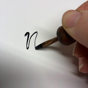 May include: A close-up of a hand using a calligraphy pen to write the lowercase letter "n" in black ink on white paper. The pen has a wooden handle and a metal nib.