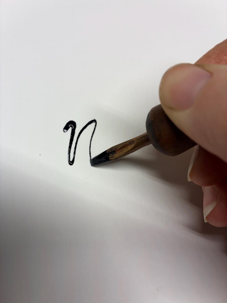 May include: A close-up of a hand using a calligraphy pen to write the lowercase letter "n" in black ink on white paper. The pen has a wooden handle and a metal nib.