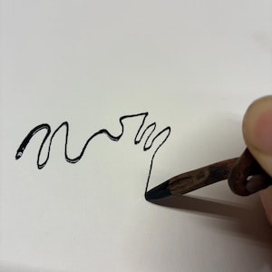 May include: A black line drawing of the word "mom" on white paper. A hand holds a pencil, the tip touching the paper to create the drawing. The pencil is brown with a black tip.