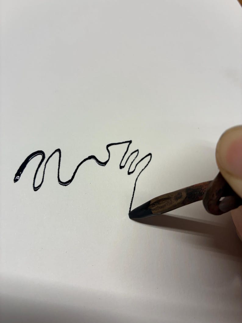 May include: A black line drawing of the word "mom" on white paper. A hand holds a pencil, the tip touching the paper to create the drawing. The pencil is brown with a black tip.