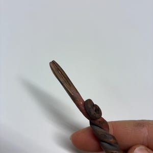 May include: A hand-forged, twisted metal tool with a pointed end. The metal has a dark, aged appearance with reddish-brown accents. The tool's design includes a spiral twist and a small, circular detail.