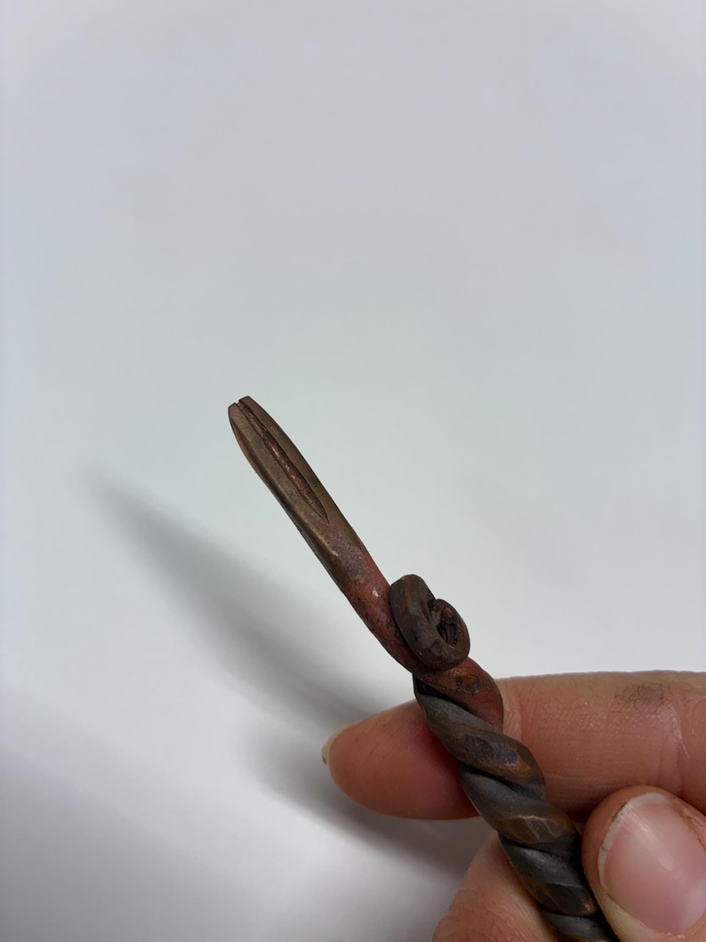 May include: A hand-forged, twisted metal tool with a pointed end. The metal has a dark, aged appearance with reddish-brown accents. The tool's design includes a spiral twist and a small, circular detail.