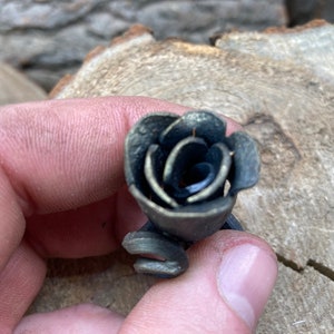 May include: A small, black metal rose with a detailed, layered design. The rose is crafted from metal and has a rustic, antique look.