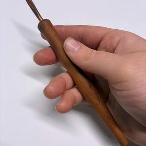 May include: A wooden crochet hook with a copper-colored tip. The hook has a smooth, polished wooden handle and a tapered design. The hook is held in a hand against a white background.