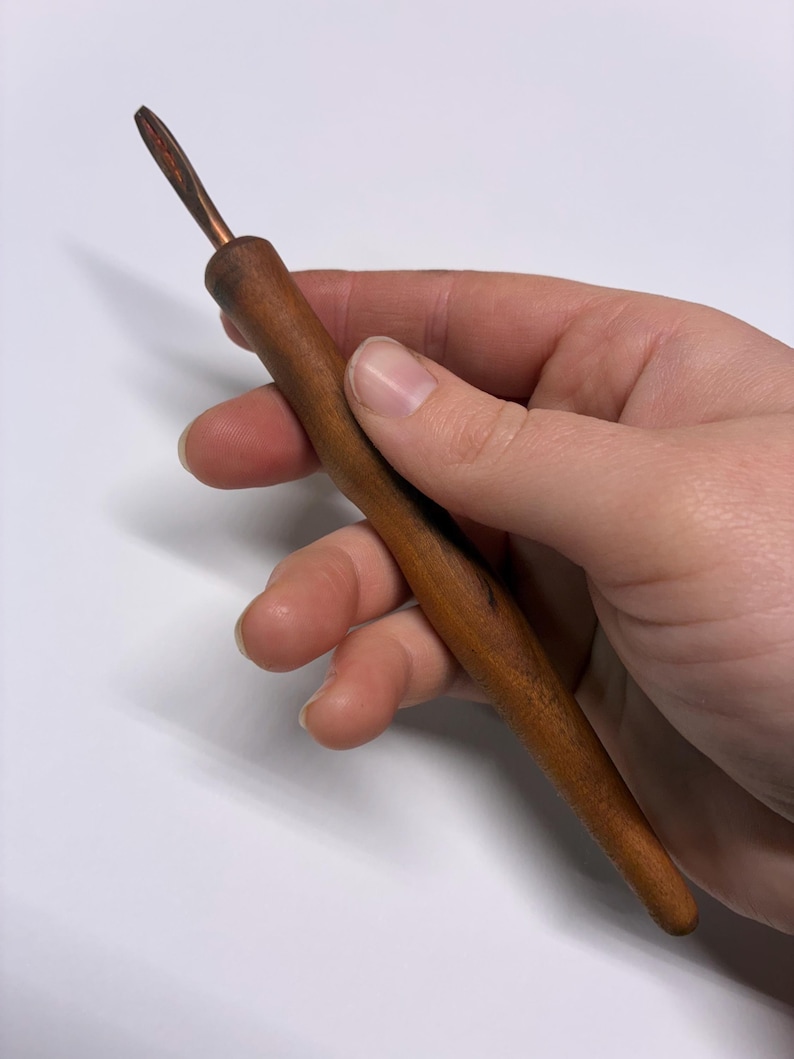 May include: A wooden crochet hook with a copper-colored tip. The hook has a smooth, polished wooden handle and a tapered design. The hook is held in a hand against a white background.