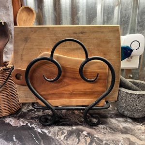 May include: A black metal cutting board holder with two wooden cutting boards. The holder has a heart shape and is made of wrought iron. The cutting boards are both rectangular and made of wood.