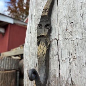 May include: Hand-forged metal hook with a rustic, weathered appearance. The hook is shaped like a stylized face with carved details, including eyes, a beard, and a star-like design. It is mounted on a weathered wooden surface.