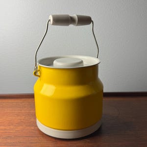 Puede incluir: Un recipiente de esmalte amarillo con base y tapa blancas. Tiene un asa plateada con un agarre de madera blanco. El recipiente es cilíndrico con una base ligeramente más ancha y una parte superior más estrecha.