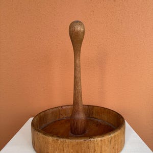Puede incluir: Un portarrollos de papel de madera, con una base circular y un poste central rematado con un botón redondeado. La madera muestra un tono cálido y natural. El soporte está colocado sobre una superficie blanca, sobre un fondo naranja.